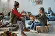 © pressmaster - Young man passing cup of tea to mature female refugee while both sitting on their sleepers among homeless people in camp