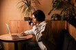 © Yuliia - She holds a coffee cup and uses her laptop. The warm tones and casual attire create a calm and inviting scene. Sitting at a wooden table, the woman combines work and leisure.
