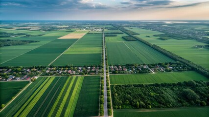 Poster - Green fields and forests create a geometric pattern across a vast agricultural landscape, bisected by a straight country road leading to a small village