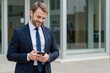 © MT - A 30-year-old Caucasian male business professional in a suit smiles while using a smartphone outside a modern office building.