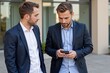 © MT - Two Professional Caucasian Men in Business Suits Engaged in Discussion Outdoors While One Uses a Smartphone in a Modern Office Environment