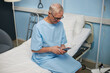 © DragonImages - Elderly man sitting on hospital bed scrolling smartphone, wearing blue hospital gown