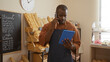 © Krakenimages.com - Young man in an apron talking on the phone while using a tablet in a bakery shop with bread and pastries displayed around him