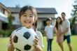 © Bambalino Studio - A young girl is holding a soccer ball and smiling. The family is outside in a grassy area