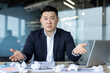 © Tetiana - Portrait of a young Asian man in a suit sitting in an office at a desk with crumpled papers scattered on it, folding his hands in frustration and looking at the camera