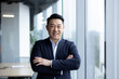 Portrait of a serious young Asian man in a business suit confidently posing for the camera in a corporate office with his arms crossed on his chest