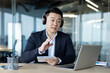 © Tetiana - Asian young serious man in headphones and suit sitting in the office in front of a laptop, talking on a video call, taking notes