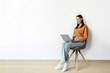 © Home-stock - Smiling woman sitting on chair with laptop computer against white wall, studying or working online with portable pc, free space