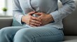 © rdkcho - Woman Experiencing Abdominal Pain or Discomfort, Holding Her Stomach, Sitting on a Couch, Healthcare, Medical, Woman's Health