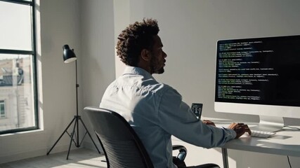 Programmer coding at desk, Focused male programmer sitting at desk and writing code on large monitor in bright modern office with smartphone and minimal furnishings by the window

