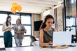 © Wavebreak Media - Focused woman working on laptop while diverse female colleagues celebrate with balloons, at office