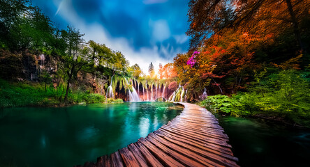 Poster - A wooden path meanders through a vibrant forest, leading towards a stunning waterfall cascading into a turquoise lake under a partly cloudy sky. Sunlight illuminates the scene, creating a tranquil and