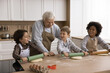 © fizkes - Little chefs. Caring old age grandpa help three multiracial grandchildren school age boys in aprons cook biscuits at home kitchen teach grandkids cooking as funny game explain how to mix roll dough