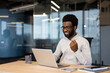 © Liubomir - An African-American man in glasses smiles and pumps his fist, celebrating success while sitting at his desk in a modern office setting.