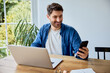 © baranq - Young man using laptop and mobile phone at home sitting at table in living room