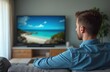 © Vadym - Man sits sofa watches TV with blue ocean view. Male wearing denim shirt looks at display screen, enjoying streaming video, online content at home. Relaxation entertainment.