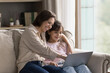 © fizkes - Digital leisure time. Friendly parent and kid young mom little girl daughter spend weekend sitting on couch with notebook use gadget for learning or entertainment. Mommy teaching child to work in app