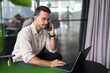 © Dorde - Young Professional Man in Neutral Shirt on Phone Working on Laptop in Modern Office