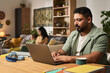 © pressmaster - Man working on laptop in a cozy living room with woman and child in background, showcasing a typical day of remote work. Various devices and gadgets are present on the desk and surroundings