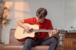© Tj - Young musician practicing acoustic guitar chords sitting on a brown leather couch at home, enjoying his hobby in a cozy living room with warm sunset light