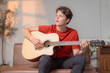 © Tj - Young man wearing a red t-shirt and black pants sits comfortably on a brown leather couch, playing an acoustic guitar and singing a heartfelt song in his cozy home