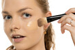 © vladimirfloyd - Close-up of young woman applying liquid foundation with makeup brush on cheek, showing smooth application process against white background.