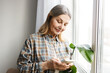 © shurkin_son - Indoor portrait of grandmother texting message in group chat of relatives sharing memes and wishing good day, standing next to window and houseplant, holding smartphone in hands, looking at screen