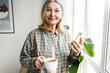 © shurkin_son - Senior lady in casual shirt with cup of coffee enjoying morning holding smartphone in hands, browsing news feed, standing in front of window and looking at camera with happy facial expression