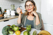 © shurkin_son - Healthy eating. Secrets of longevity. Senior lady in glasses enjoying breakfast at home, sitting at kitchen table with spoon in hand, having home-made green smoothie prepared form organic fruits