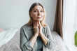 © shurkin_son - Indoor portrait of worried senior woman siting on bed in bedroom examining her thyroid, touching her throat, looking aside with anxiety, trying to find symptoms of severe endocrine disease