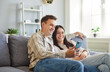© Studio Romantic - Young married couple with mobile phone sitting on sofa together. Happy man and woman relaxing on couch at home, looking at smartphone, smiling, watching video, shopping online, or using banking app
