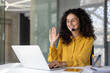 © Liubomir - A cheerful woman with curly hair, wearing a yellow shirt and headset, waves hello during a virtual meeting on her laptop