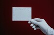 © Elena - A gloved hand elegantly holding a blank white card against a luxurious red patterned background. Ideal for branding, invitations, business, or vintage themes
