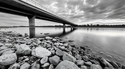 Aufkleber - Black and white photograph of a bridge over calm water with pebbled riverbank on a cloudy day : Generative AI