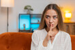 © Andrii Iemelianenko - Shh be quiet please, silent. Portrait of young woman presses index finger to lips makes silence hush gesture sign do not tells gossip secret at home apartment. Pretty girl sitting on couch. Lifestyle