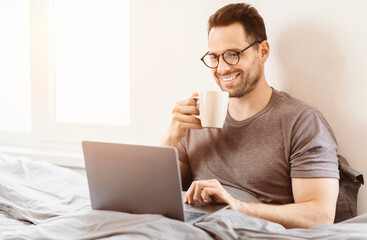 Aufkleber - Happy Man Using Laptop Browsing Internet Relaxing Having Morning Coffee Sitting In Bed At Home On Weekend.