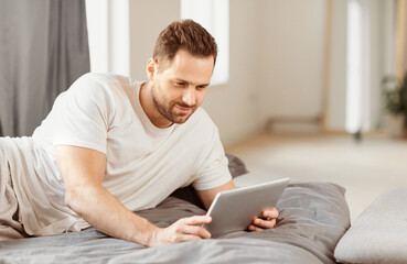 Aufkleber - Man In Casual Clothes Using Digital Tablet Lying In Bed Relaxing Watching Movie Online In Bedroom At Home. Guy With Laptop Watches Film. Gadget, Technologies, Modern Lifestyle. Weekend Leisure Concept