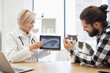 © sofiko14 - Female veterinarian in white coat showing x-ray image of a cat to male owner. Scene depicts cat healthcare and consultation in veterinary clinic. Male, adult, wearing black plaid shirt.