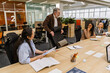 © Drobot Dean - A middle-aged White male boss in his late 40s, smiling happily as he looks over a diverse group of his female employees, while standing next to their desk in an office room
