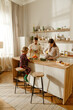 © Drobot Dean - A White family prepares breakfast together in a kitchen. A school-aged son sits on a stool at a wooden counter, while the adult father and mother prepare food. Open shelves hold kitchen items.
