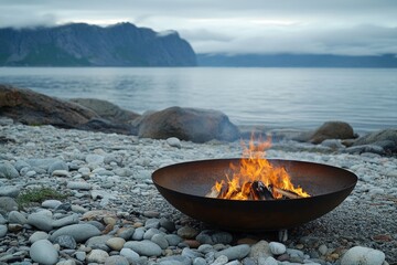 Poster - Campfire burns brightly on rocky shore with mountains in the background during overcast evening
