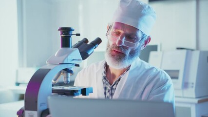 Wall Mural - bearded mature medical scientist in white coat and glasses using microscope and laptop, conducting r