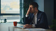 © HudHudPro - Stressed businessman in a suit sits at a desk, rubbing his forehead in frustration. A laptop, documents, and a coffee cup are on the table. Concept of workplace stress and business challenges