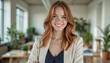 © f_bossa - Portrait headshot photo of diverse friendly people: A confident woman with auburn hair smiles warmly while standing in a bright, modern office space.