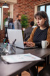 © DC Studio - Portrait of female manager focused on her laptop, preparing business project plan in brick wall office. Professional woman sits at desk with coffee, browsing for marketing strategies on digital device