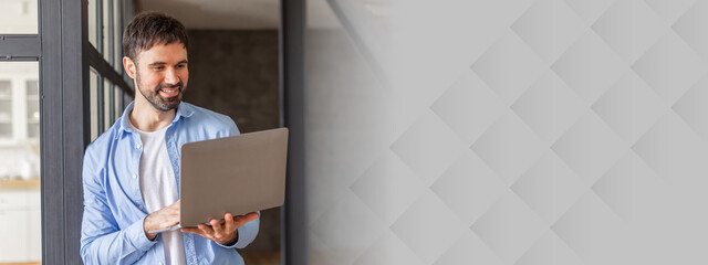 Poster - A man stands in a modern office space, smiling as he uses his laptop. He is dressed in a casual blue shirt and is leaning against a glass wall. He has short, dark hair and a beard.