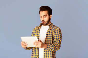 Poster - Shocked Indian man staring at tablet screen with open mouth on violet background, panorama