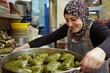 © ManusiaIkan - A woman in a hijab wraps stuffed grape leaves with precision, placing them in a large pot. She smiles as she prepares the dish her family loves
