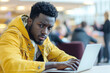© whitestorm - Stylish young African American man in yellow jacket intently working on a laptop, with a focused gaze and a modern aesthetic in a bustling co-working space.