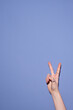 © Seventyfour - Close-up of hand displaying peace sign against plain blue background. Fingers forming iconic V-shape symbolizing peace and victory